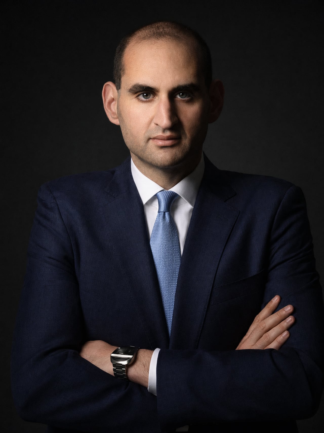 Jason Turkish in a navy suit with arms crossed, lit by a single hard key light against a dark background.